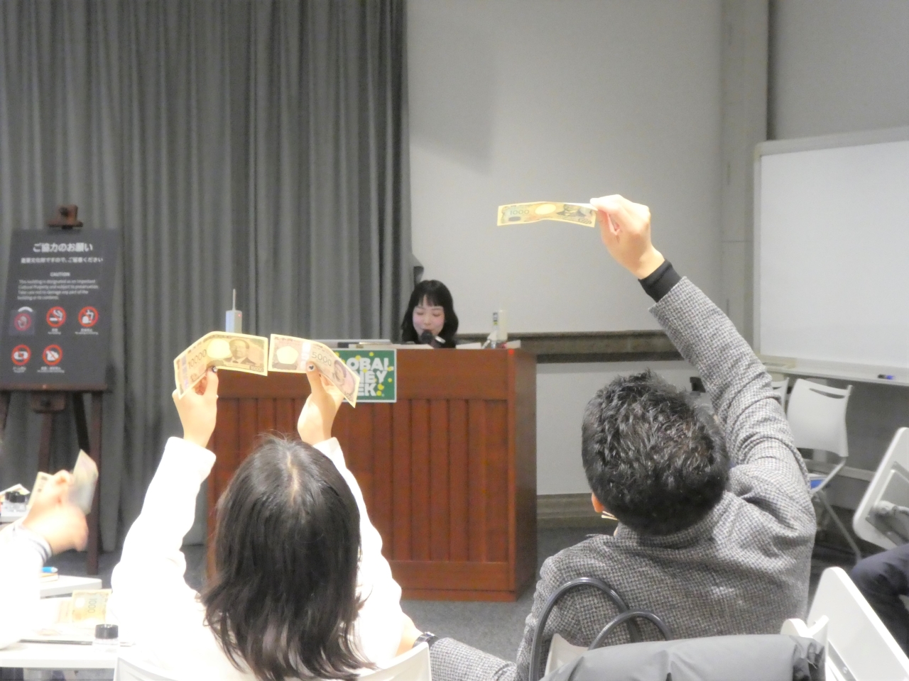 講師1人が、参加者にお札の偽造防止技術について説明し、親子(父と娘)2人が、お札を透かしてすき入れ模様を確認している様子