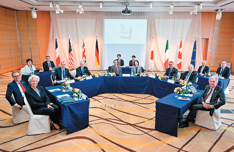 2023年G7中央銀行セッション参加者が会議テーブルに着席している写真。参加者は一斉に画面前方を見ている。会場後方には参加国の国旗が掲げられている。