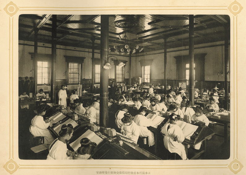 明治期の業務風景の写真。室内に多くの机が並び、数十名の職員が業務を行う様子が収められている。