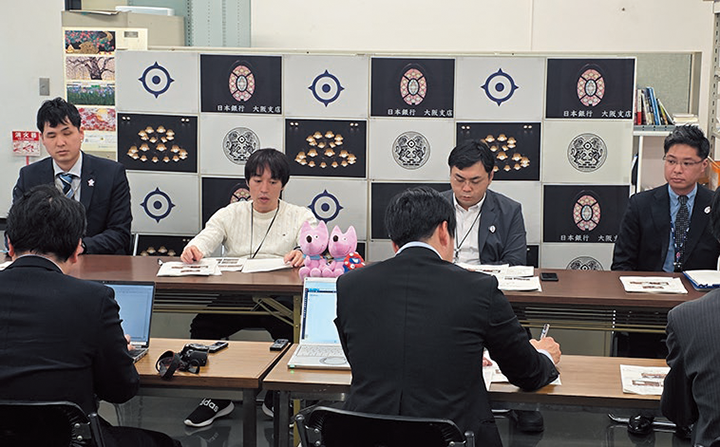 調査レポートの記者向けレクチャーの模様を撮影した写真。4人の出席者が並んで着席し、左から2番目の出席者が記者に対して説明を行っている。