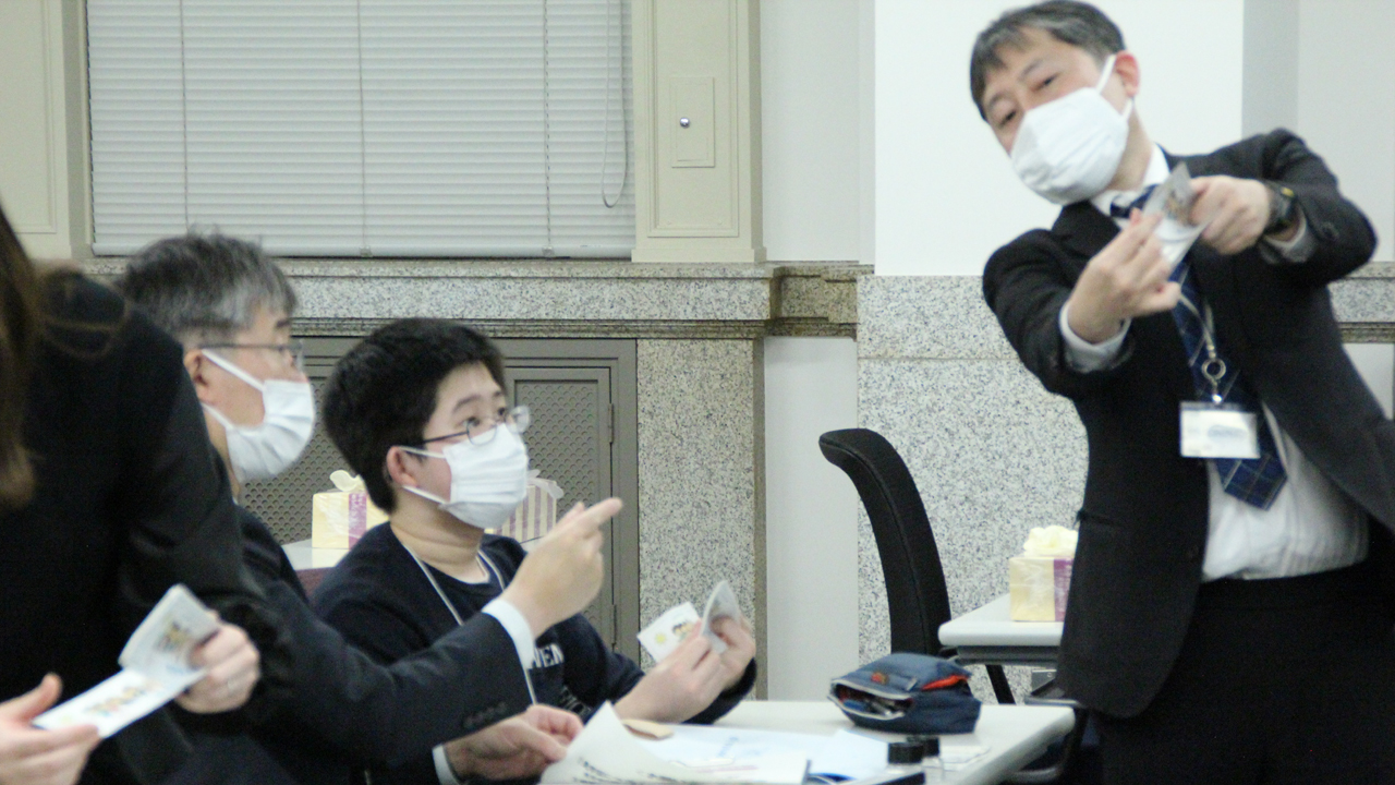 会場内にいる参加者2人が、講師1人によるお札の数え方の実演を見ている様子