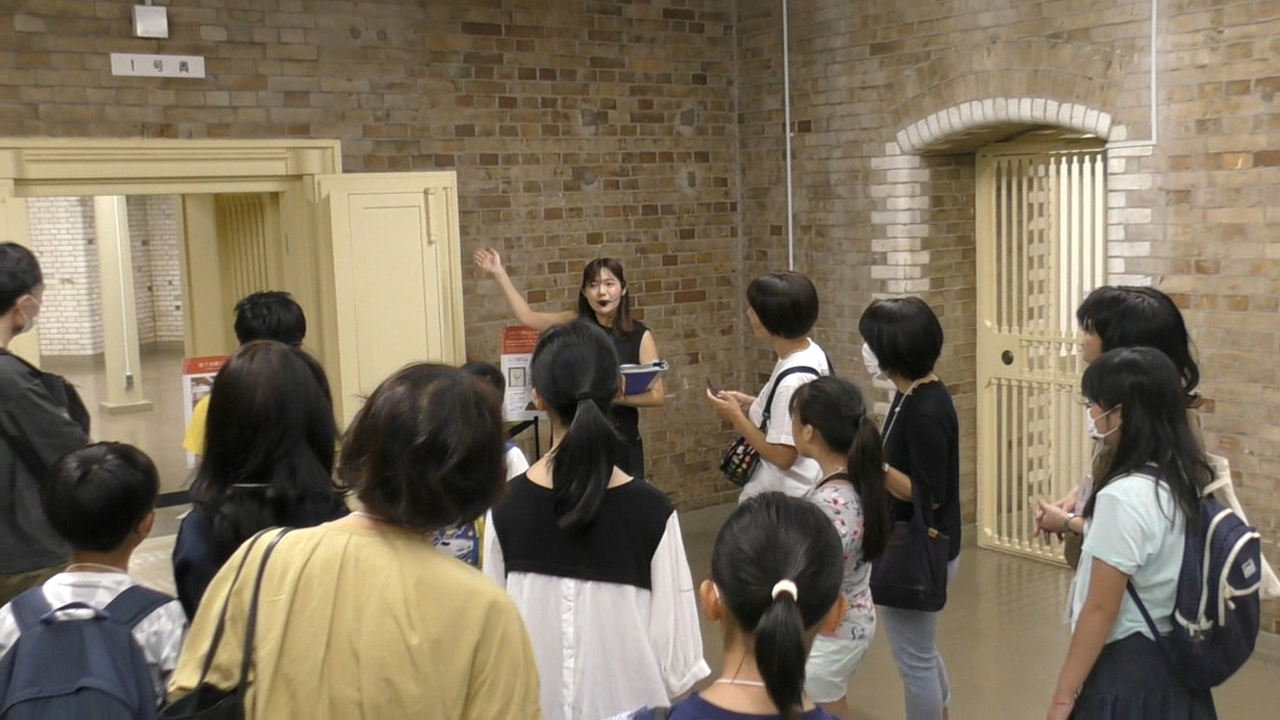 地下金庫内にいる案内者1人が、参加者13人に対して金庫扉の解説をしている様子