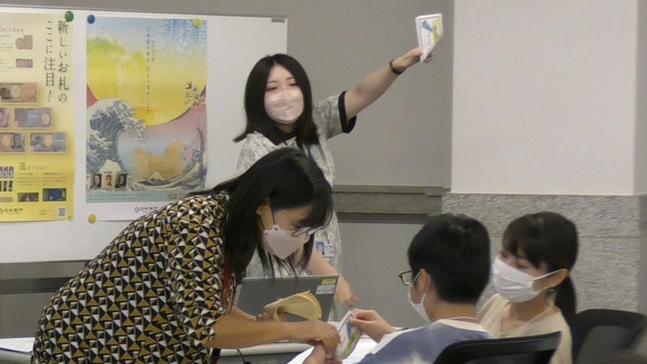 会場内にいる講師2人のうち1人がお札の数え方を実演している様子と、もう1人の講師が参加者2人に対してお札のめくり方を教えている様子