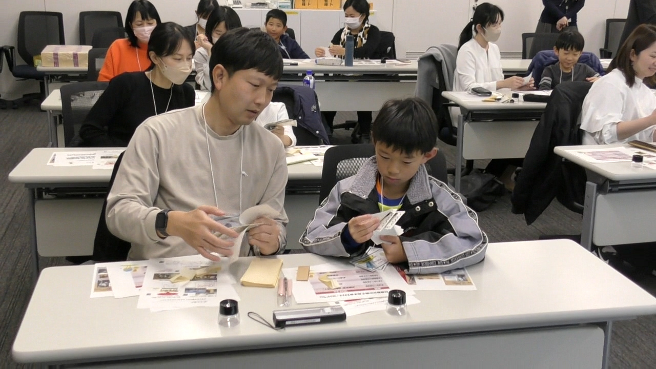 参加者11人がお札の数え方についての講義を受けている中で、参加者2名が会場前方席でお札を数えている様子