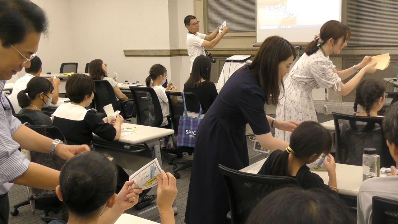 案内者4人が、参加者20人のテーブルを周り、お札の数え方を教えている様子