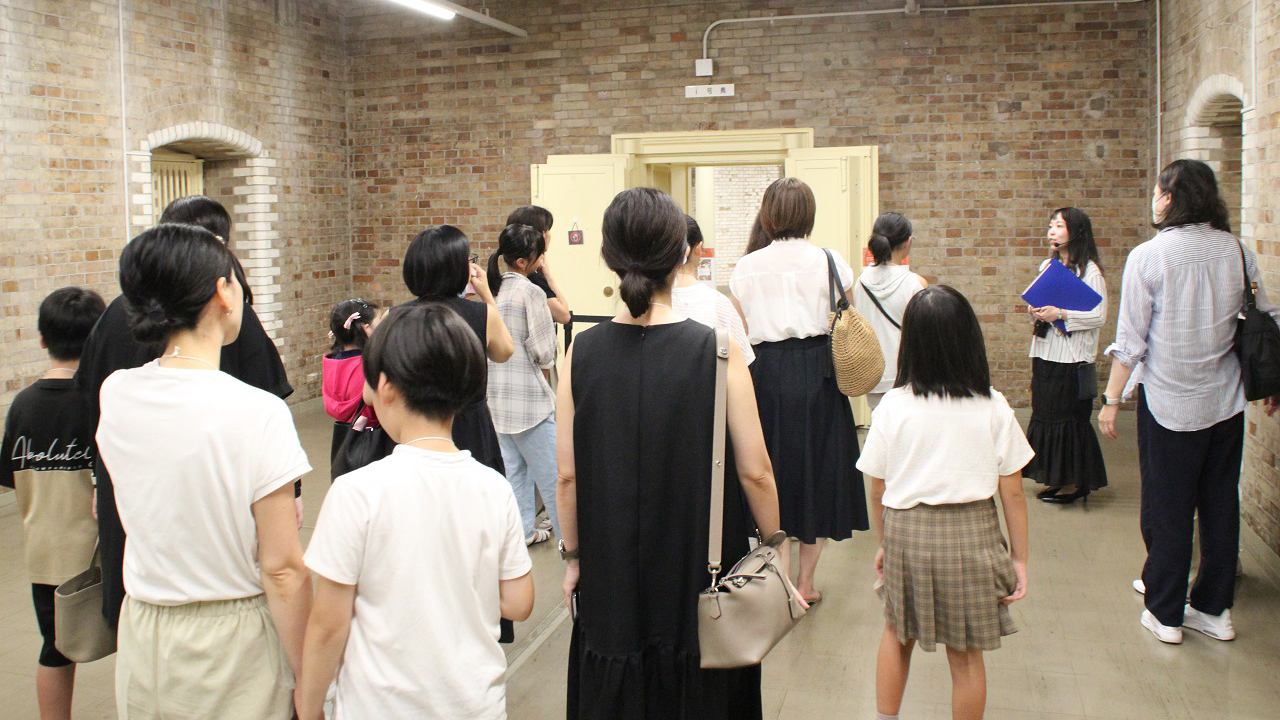 案内者1人が、参加者20人に対して地下金庫の扉を解説している様子
