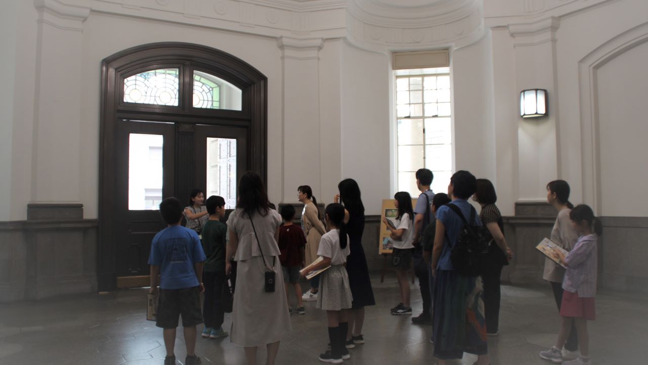 ガイド1人が、参加者20人に正面玄関内にて、建物の説明をしている様子