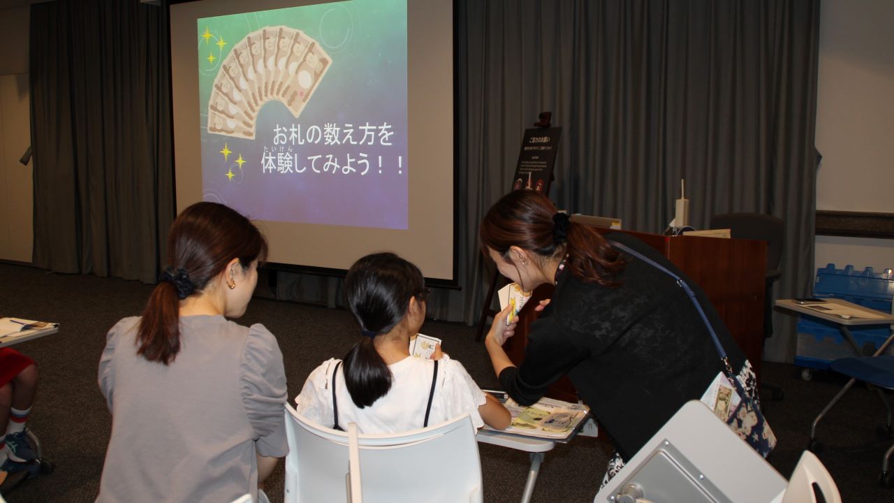 講師1人が、親子（母と娘）2人にお札の数え方を教えている様子