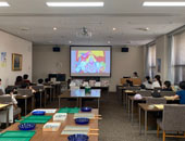 参加者が会場前方のモニターに投影されたDVD（子供向け学習用教材）を視聴している写真