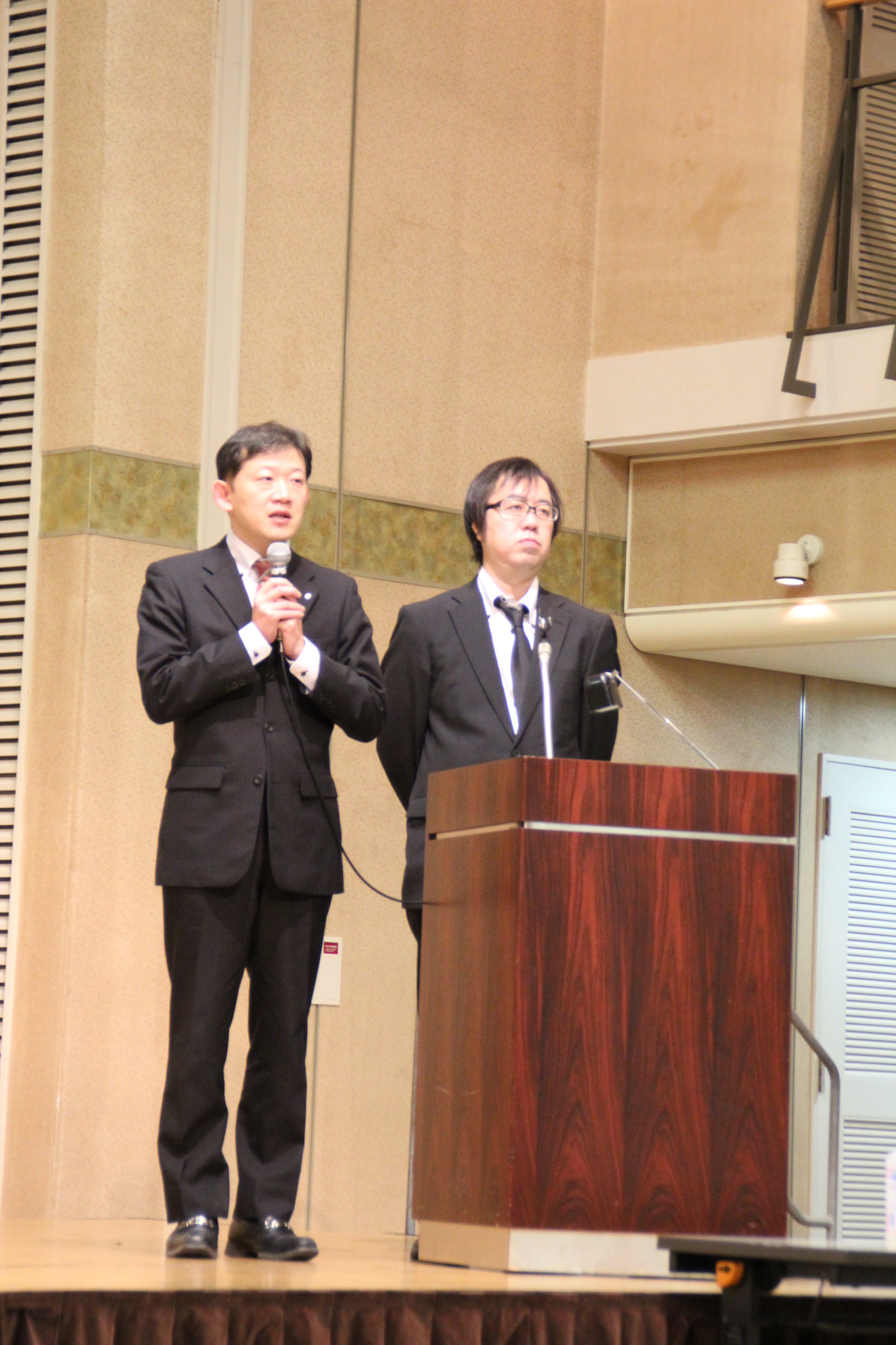 マイクを持って話している講演者とその隣に立つ別の講演者の写真